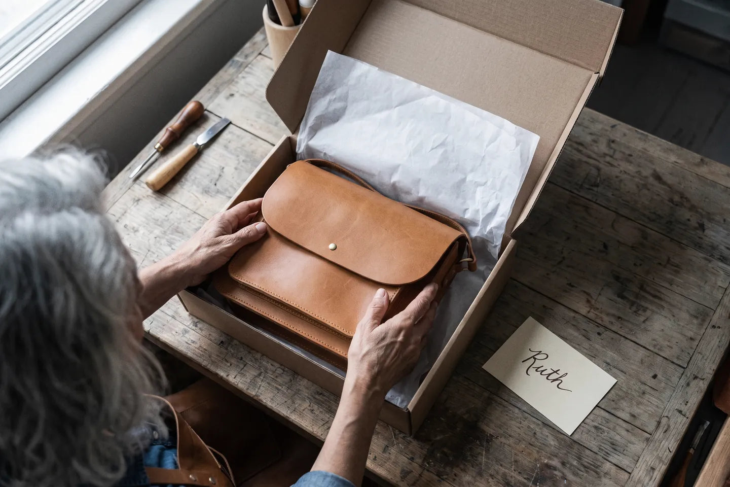 Rose&rsquo;s hands laying a finished bag into a tissue-lined box, a handwritten name card beside it.