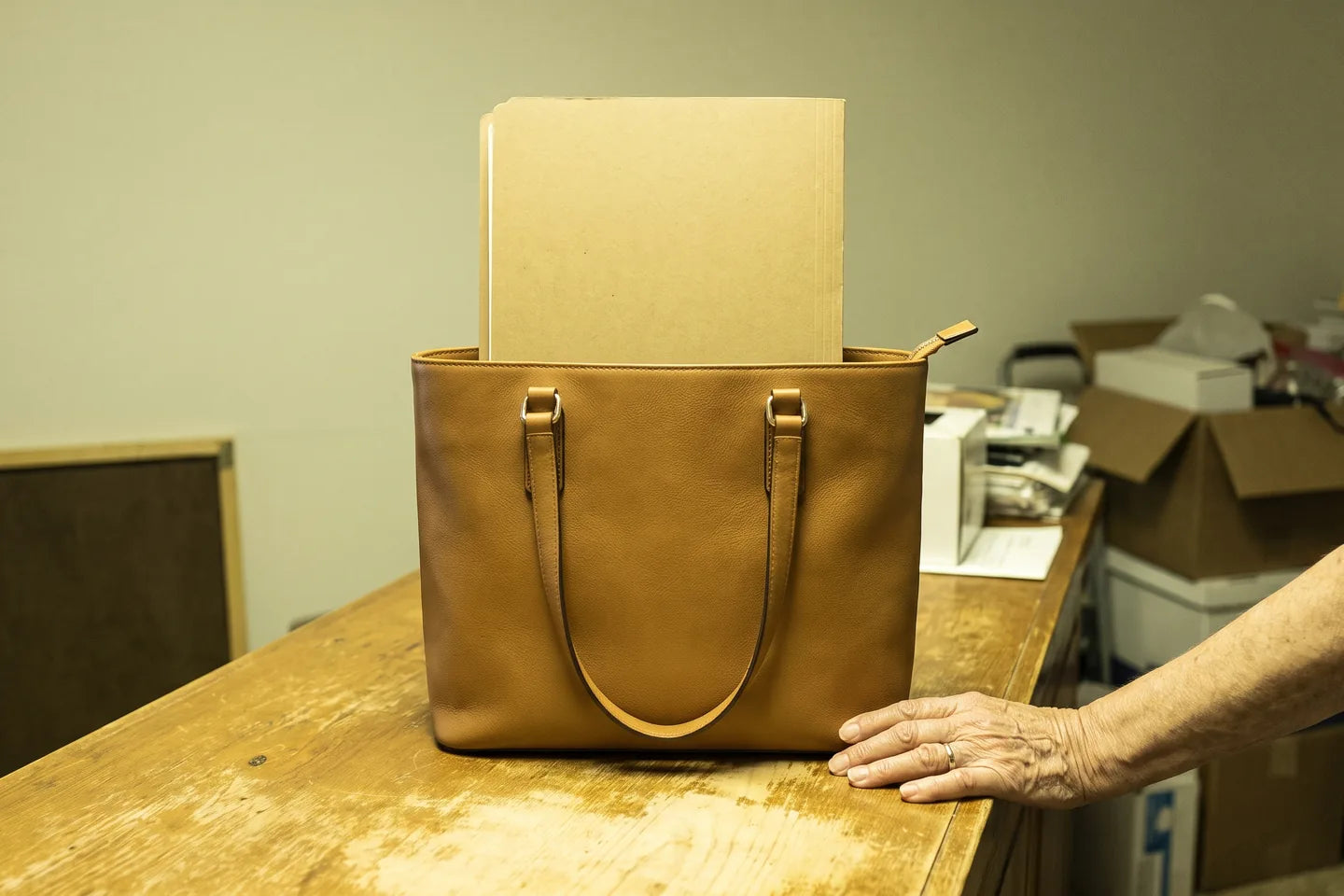 A deep leather tote standing upright and unaided on a bare counter with a blank folder standing square inside it.