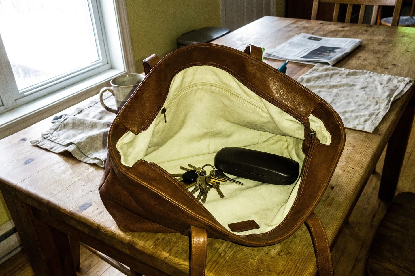 A leather bag open on a pale table, the pale interior clearly visible, keys and a glasses case in plain sight against the light lining.